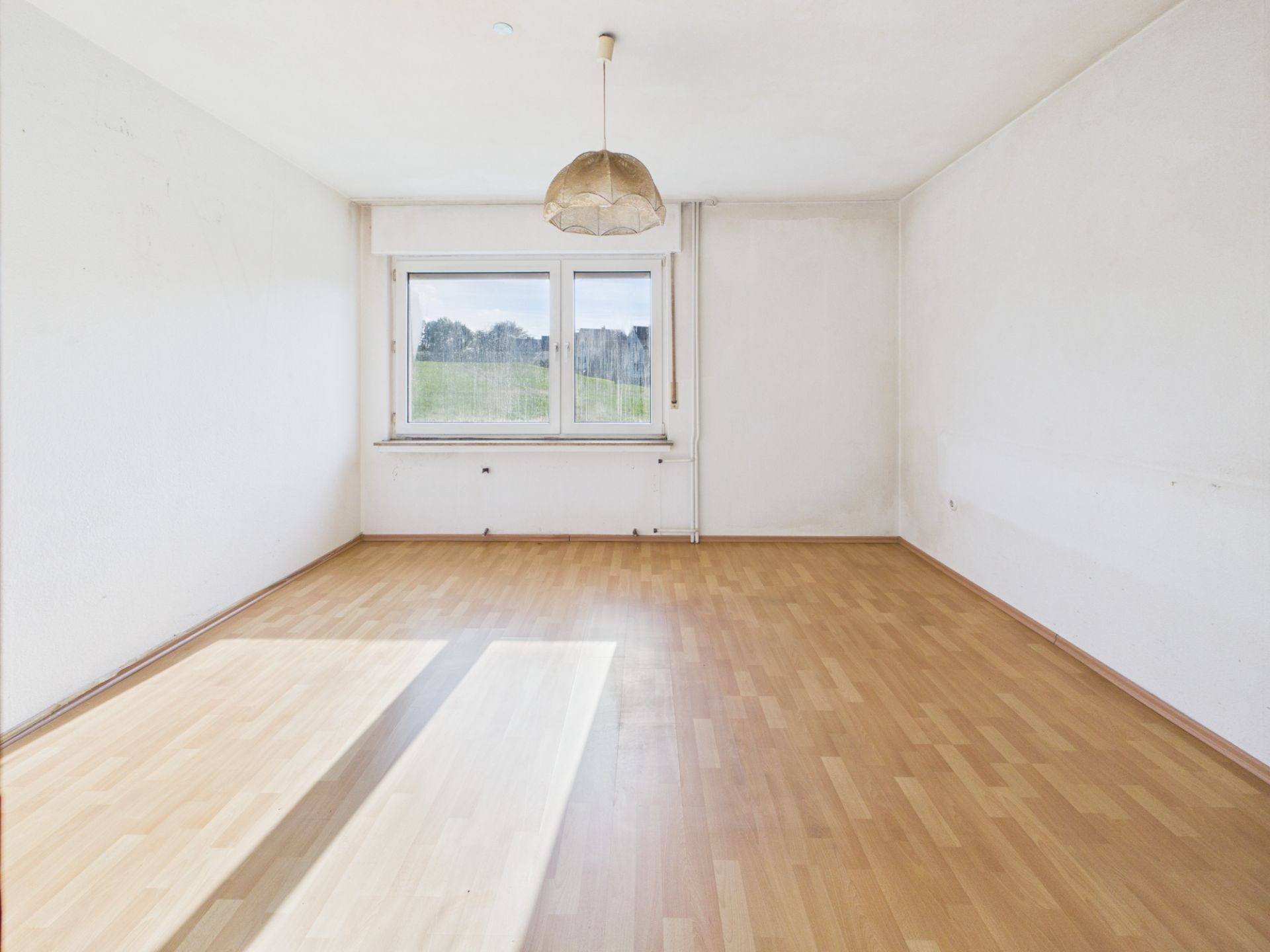 Leeres Zimmer mit Holzboden, weiß gestrichenen Wänden und großem Fenster.