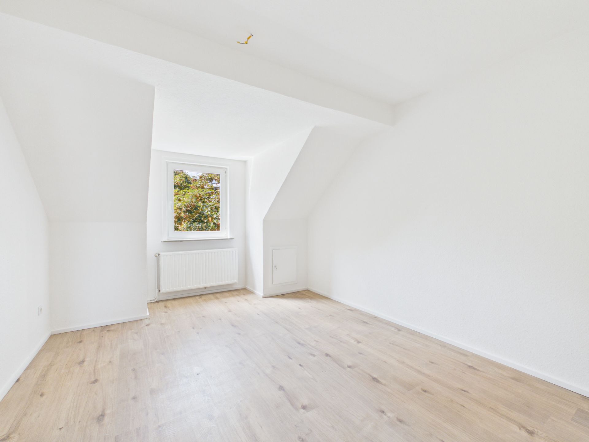 Helles, leeres Zimmer mit weiß gestrichenen Wänden, Holzfußboden und einem Fenster mit Heizung darunter.