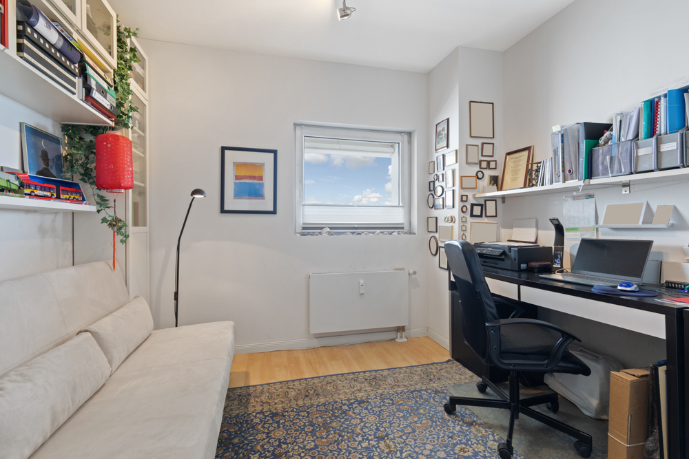 Helles Arbeitszimmer mit Schreibtisch, Stuhl, Sofa, Regalen und Fenster.