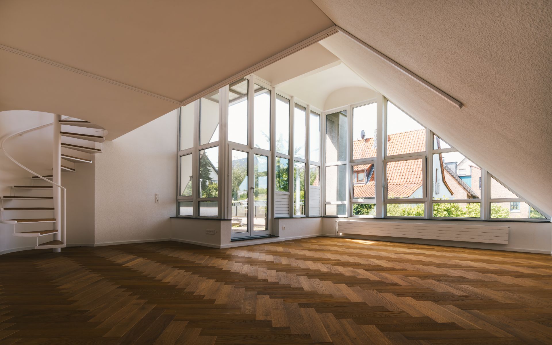 Großer, heller Raum mit schräger Decke, Parkettboden, Glasfront und Wendeltreppe.