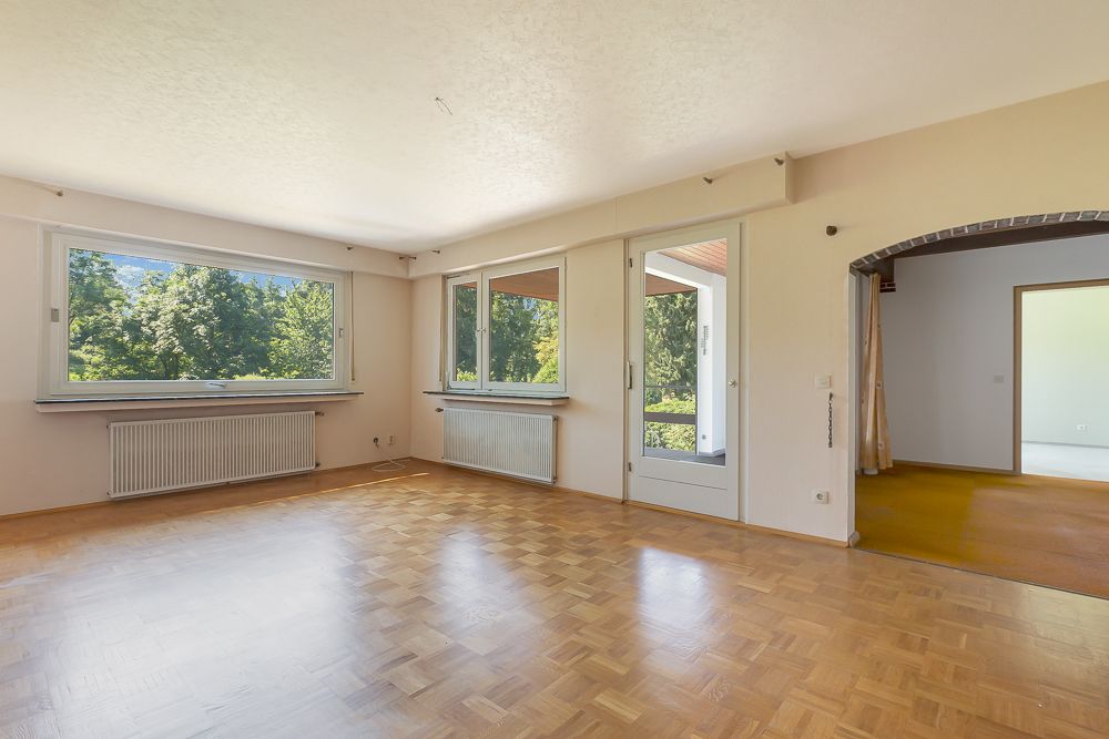 Helles Wohnzimmer mit Parkettboden, großen Fenstern und Zugang zu einem anderen Raum durch einen Rundbogen.