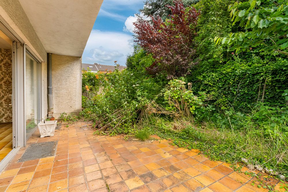Terrasse mit braunen Fliesen und angrenzendem, überwuchertem Garten an einem Wohnhaus.