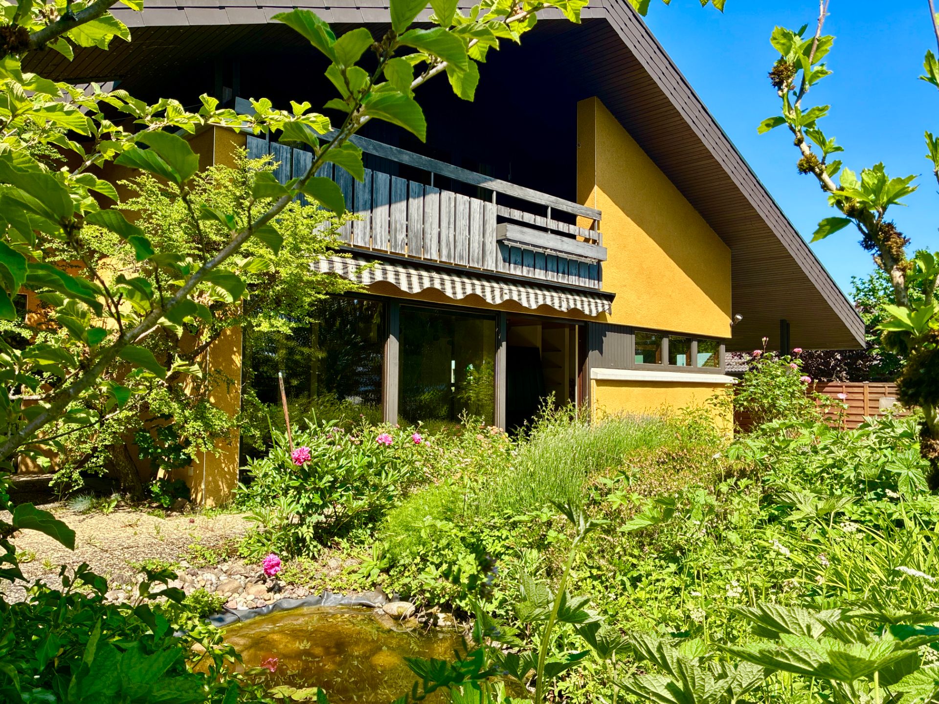 Haus mit großem Balkon, gelber Fassade und üppigem Garten mit Teich im Vordergrund.