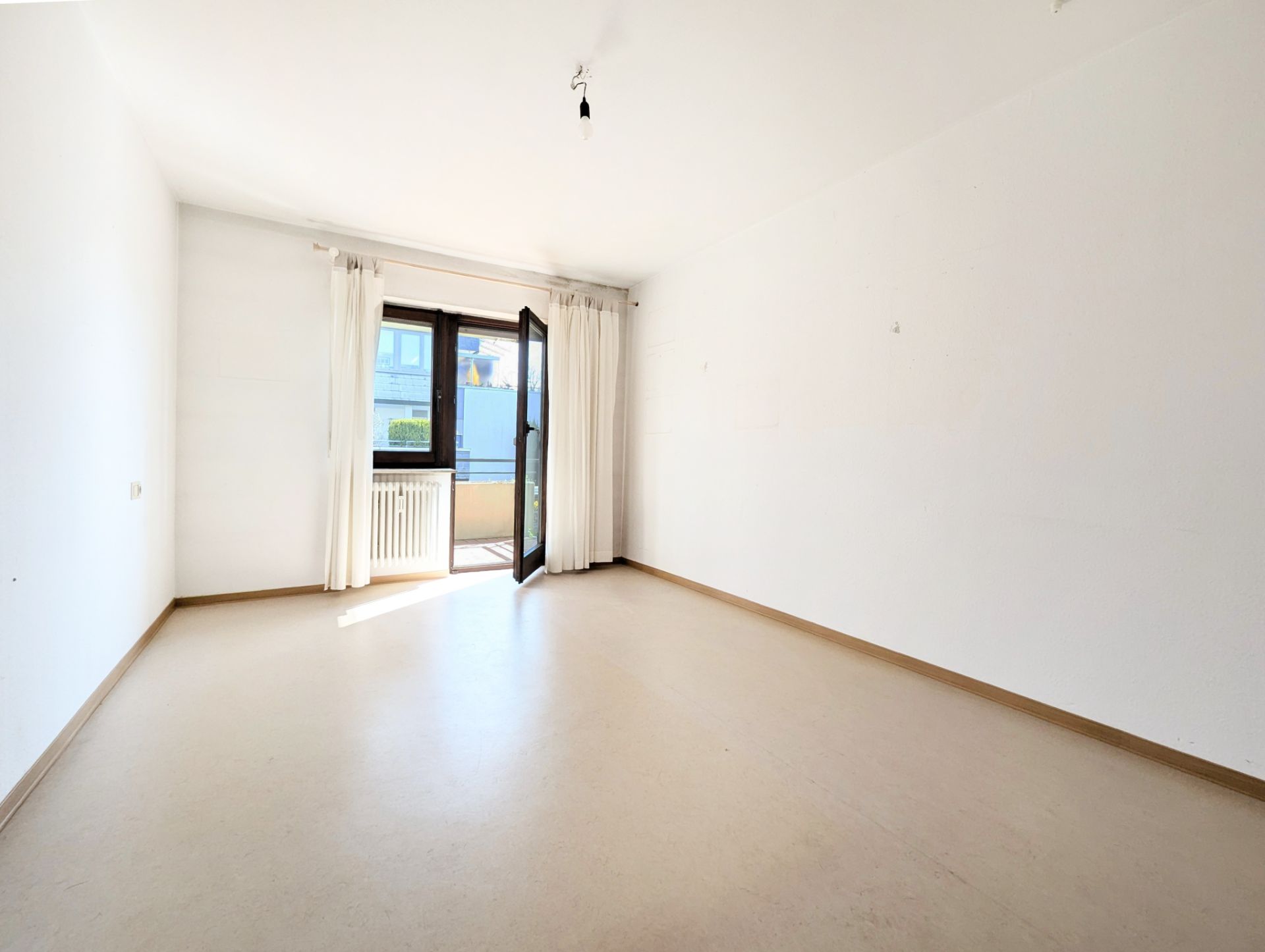 Helles, leeres Zimmer mit offenem Fenster und weißen Vorhängen, Zugang zu einem kleinen Balkon.
