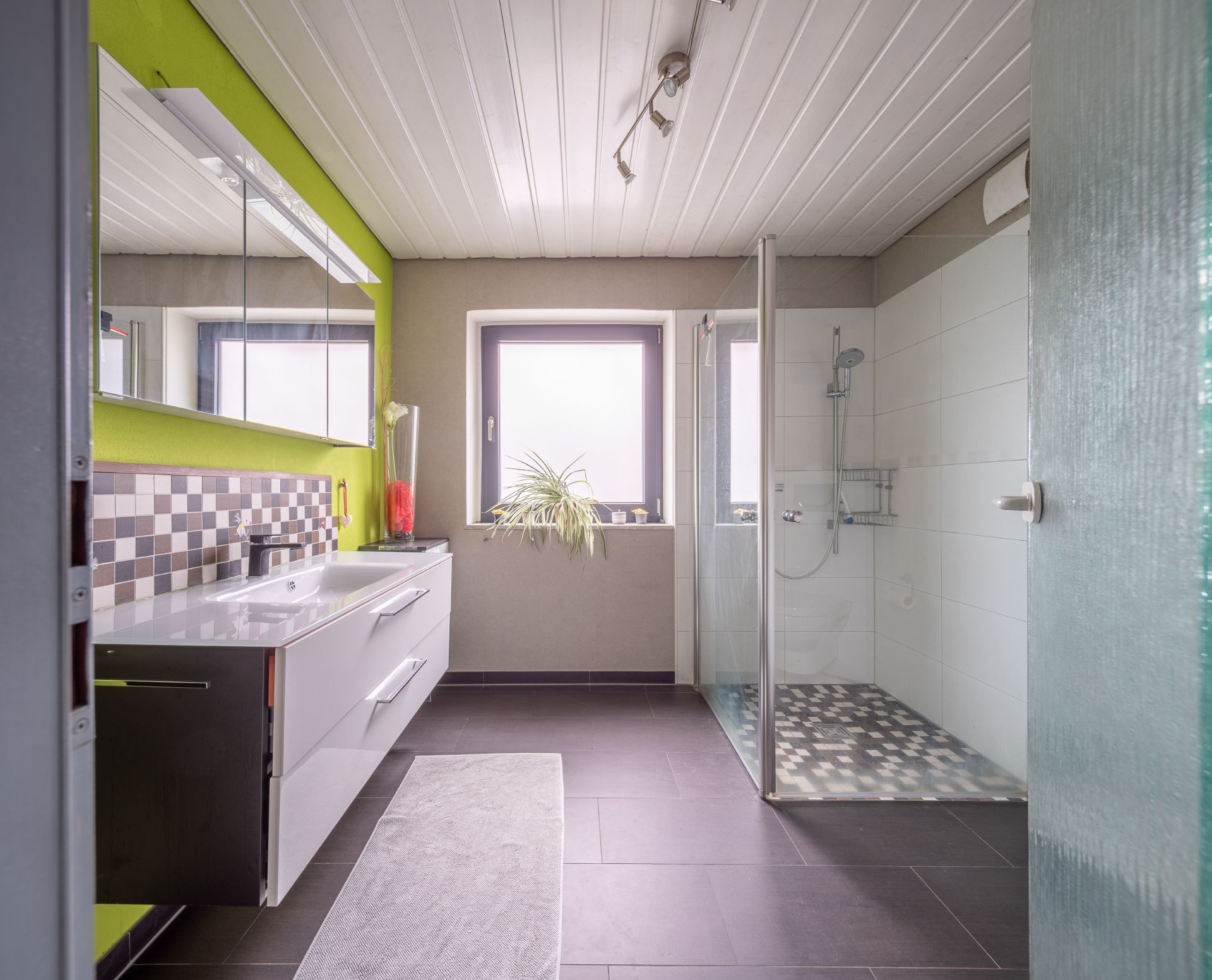Modernes Badezimmer mit großer Dusche, Waschbecken, Spiegel und Fenster, helle Fliesen und grüne Wand.