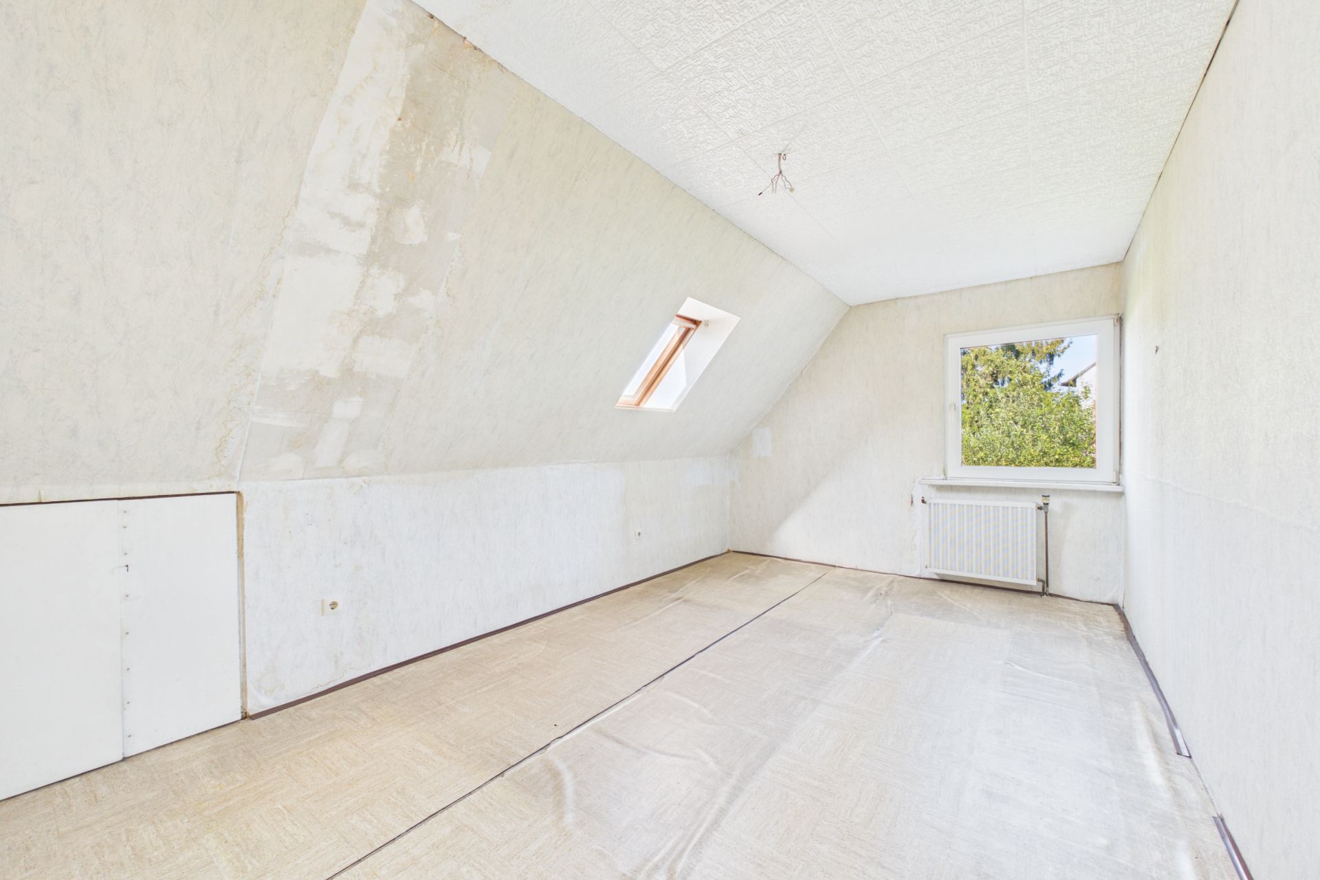 Helles, leeres Zimmer mit schräger Decke, Dachfenster und normalem Fenster mit Blick ins Grüne.