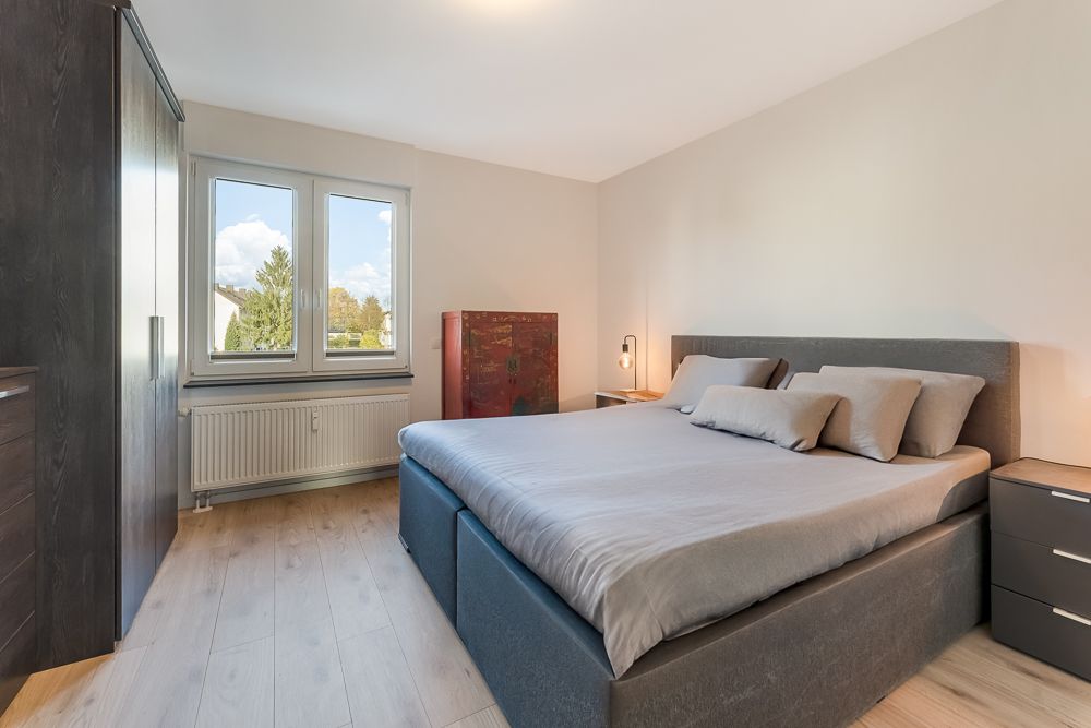 Modernes Schlafzimmer mit großem Bett, zwei Fenstern und Kleiderschrank auf hellem Holzboden.