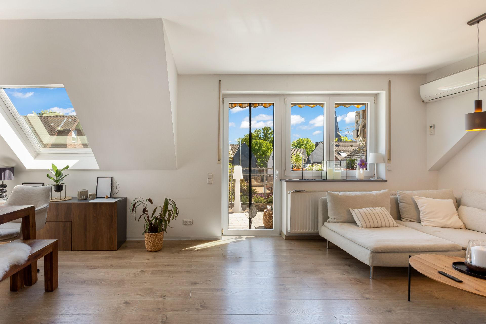 Helles Wohnzimmer mit Ecksofa, Holzboden, Pflanzen und Zugang zum Balkon.