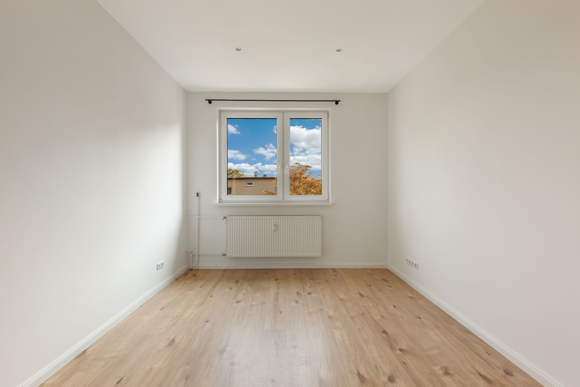 Heller leerer Raum mit Holzfußboden, weißer Wand und Fenster mit Blick nach draußen.