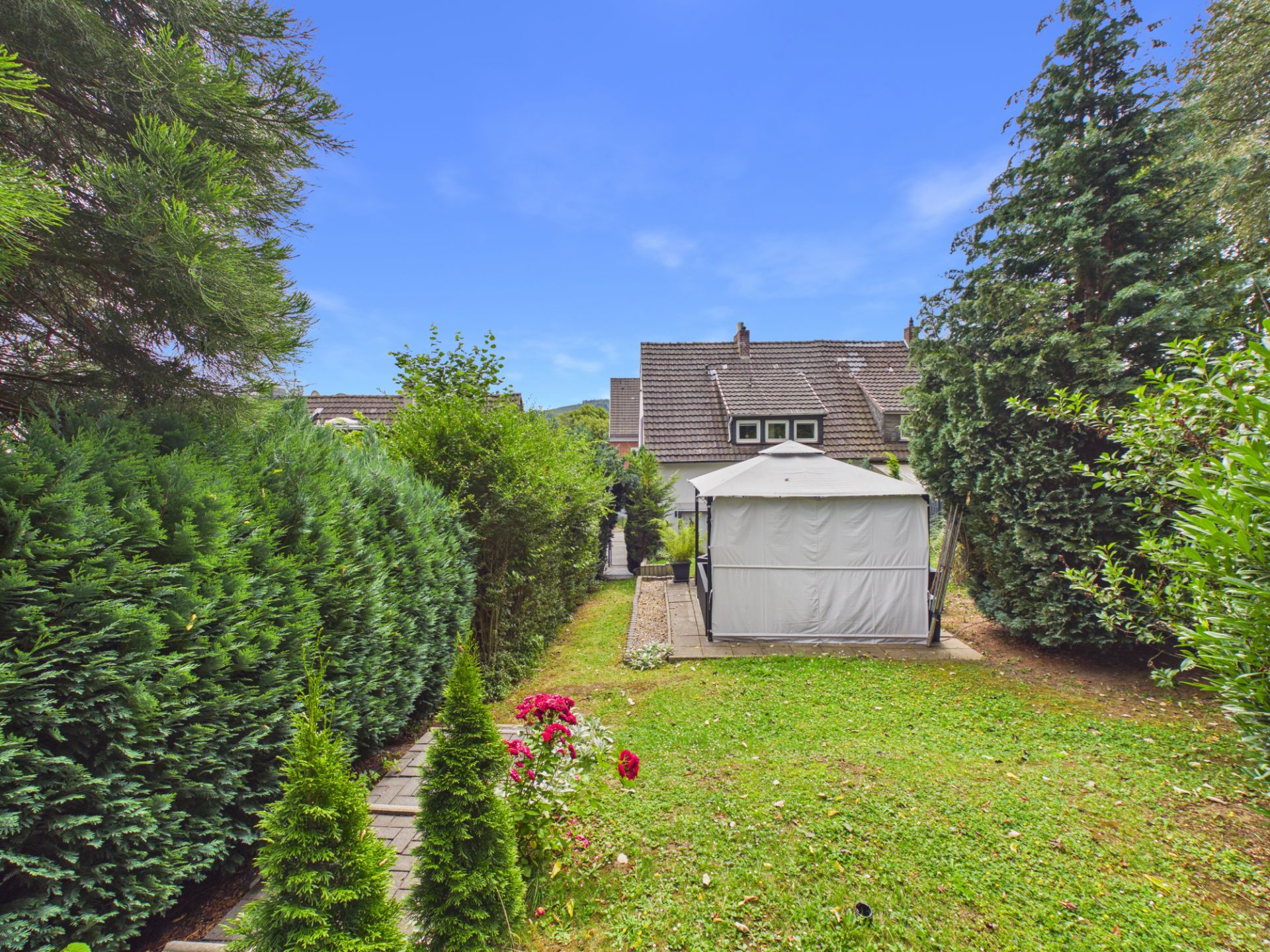 Garten mit grünem Rasen, Zeltpavillon, Blumen und hohen Büschen, im Hintergrund ein Wohnhaus.