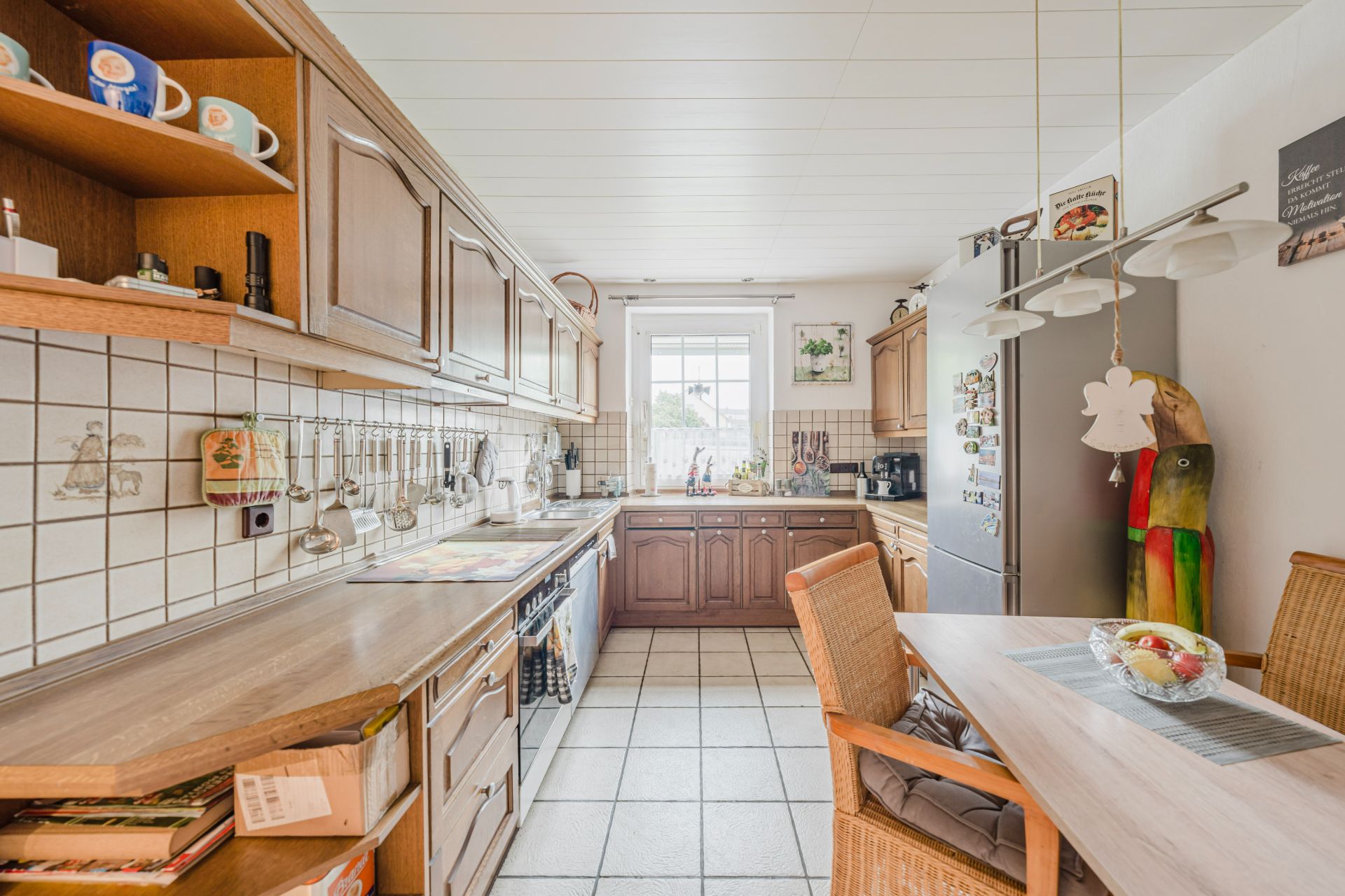Helle Küche mit Holzschränken, Esstisch, Kühlschrank und Fenster am Ende des Raums.