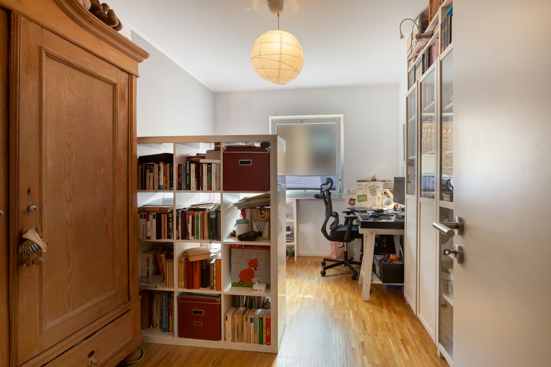 Helles Arbeitszimmer mit Bücherregal, Schreibtisch, Bürostuhl und großem Holzschrank.