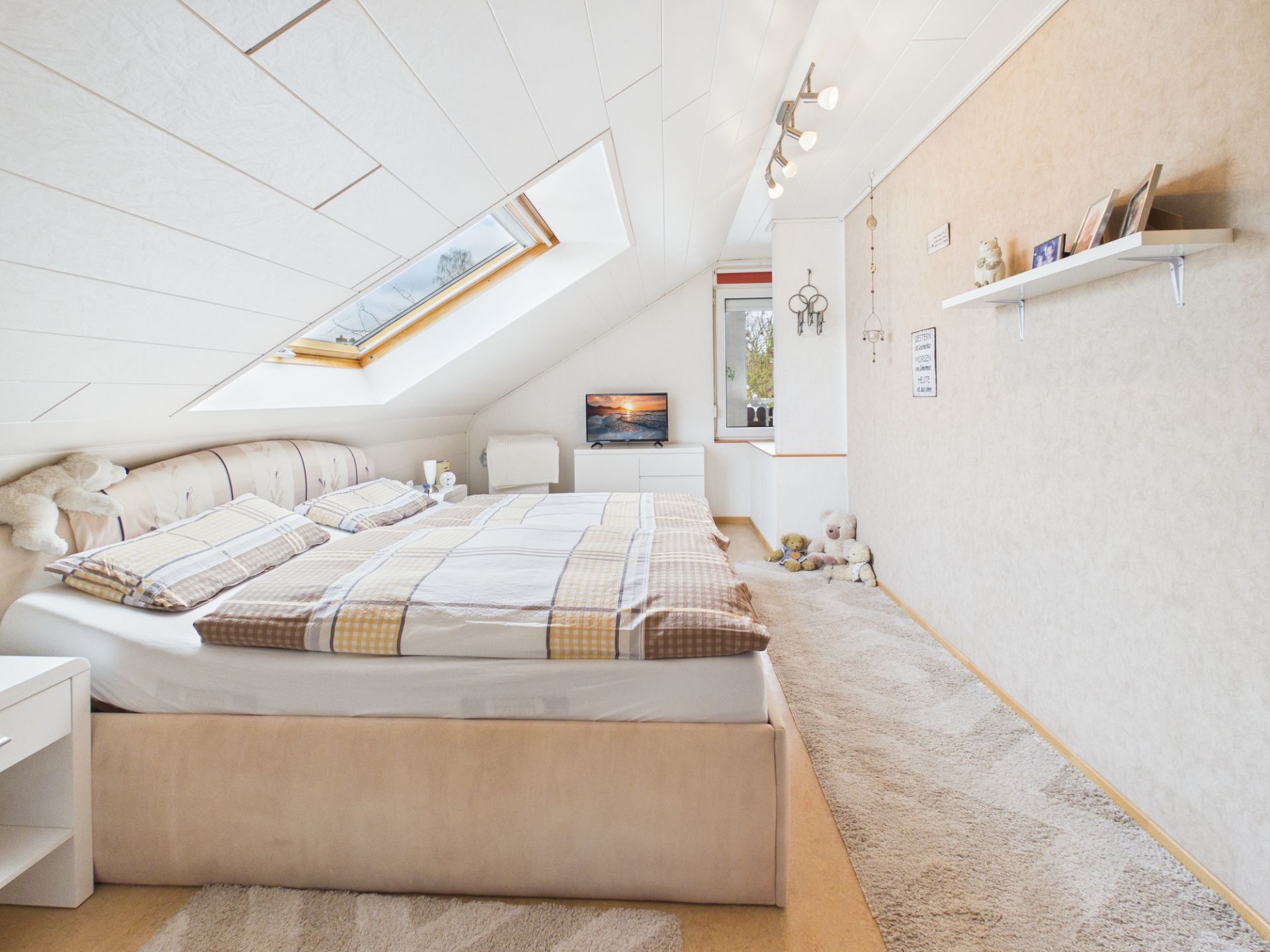 Helles Schlafzimmer mit Dachfenster, Doppelbett und Fernseher unter einer Dachschräge.