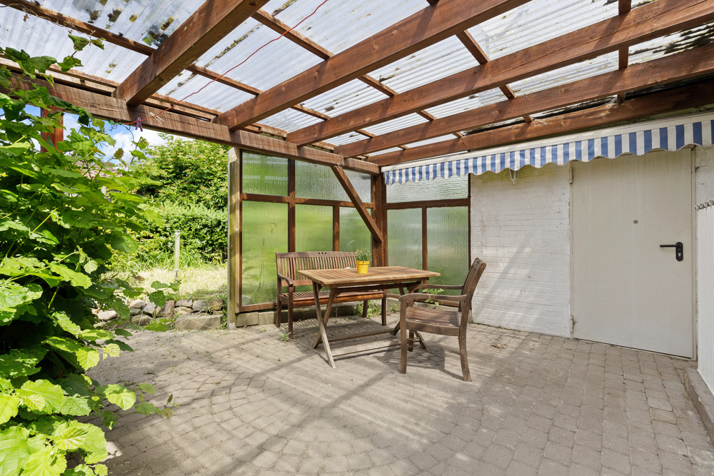 Überdachte Terrasse mit Holztisch, zwei Stühlen und Bank auf Steinboden neben einer weißen Tür.