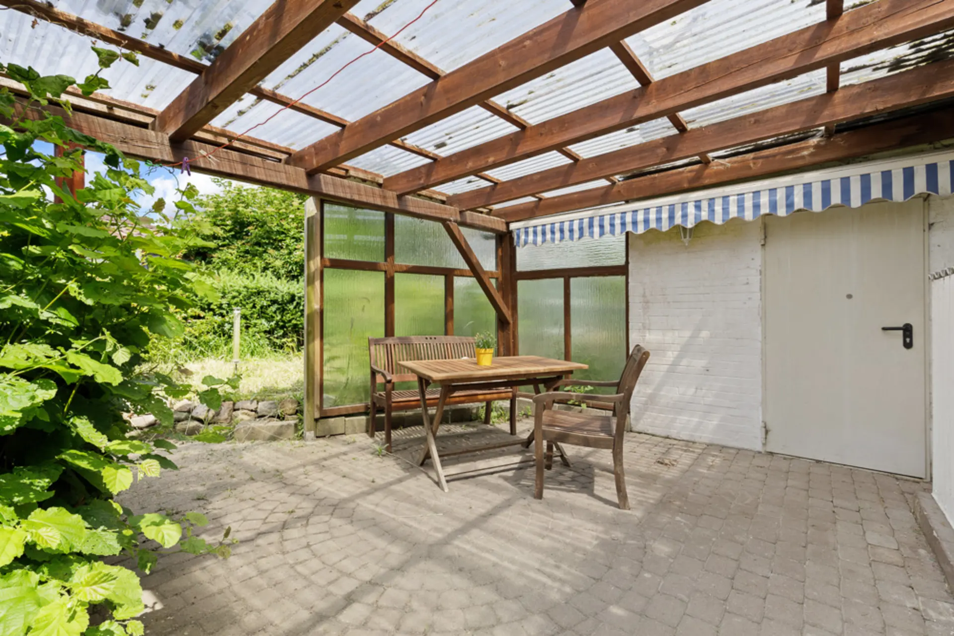 Überdachte Terrasse mit Holztisch, zwei Stühlen und Bank auf Steinboden neben einer weißen Tür.