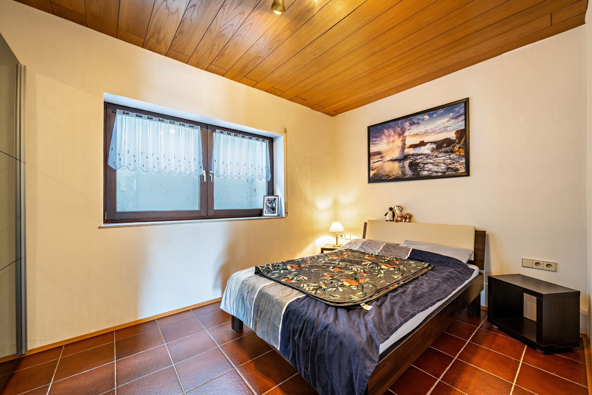 Schlafzimmer mit Doppelbett, Nachttisch, großem Fenster und Wandbild, Holzdecke und Fliesenboden.