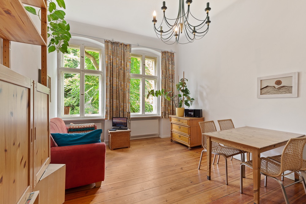 Helles Wohnzimmer mit Holzboden, rotem Sofa, Esstisch, Pflanzen und großen Fenstern.