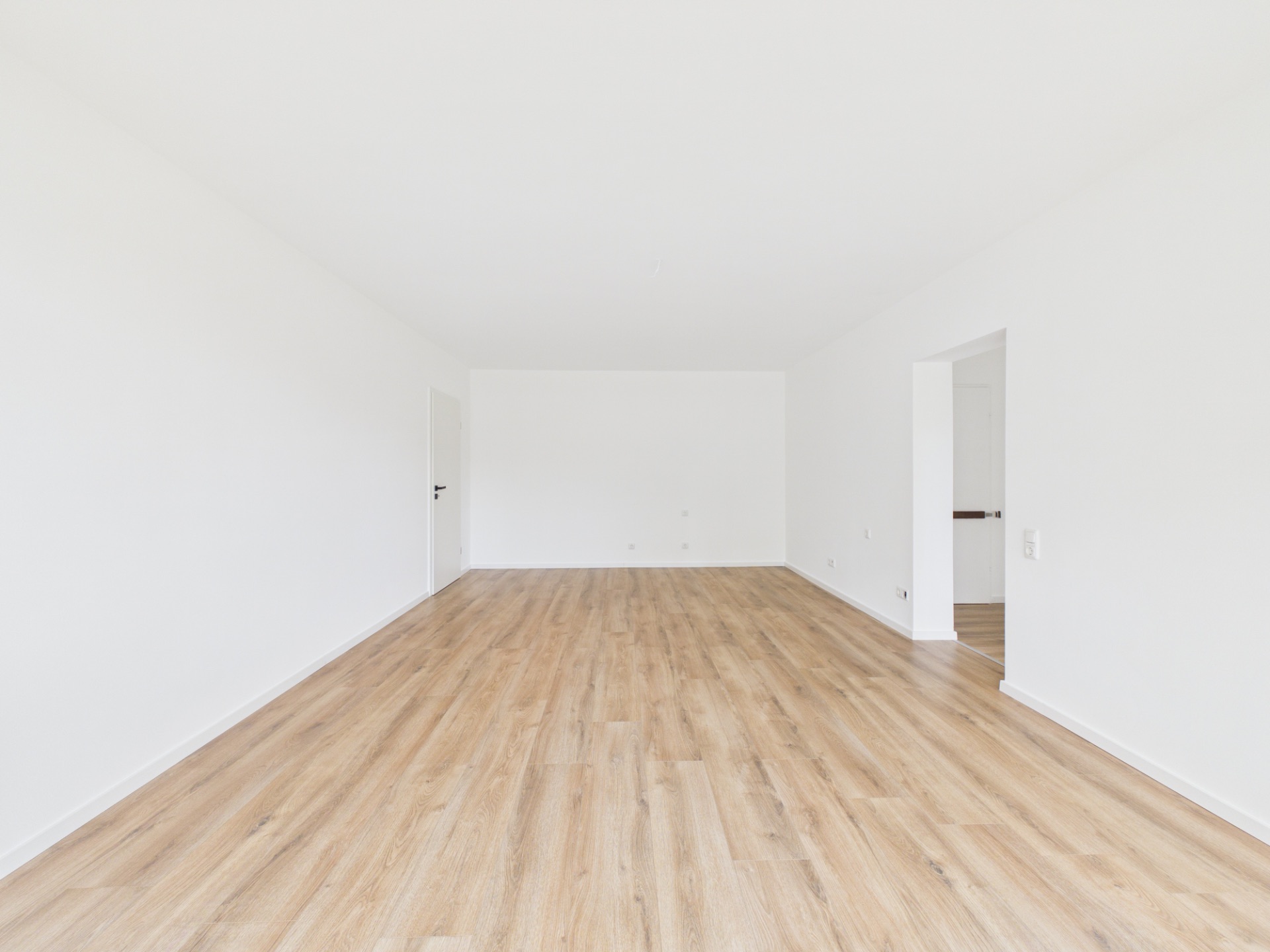 Leerer, heller Raum mit Holzfußboden und weißen Wänden, zwei Türen an den Seiten.