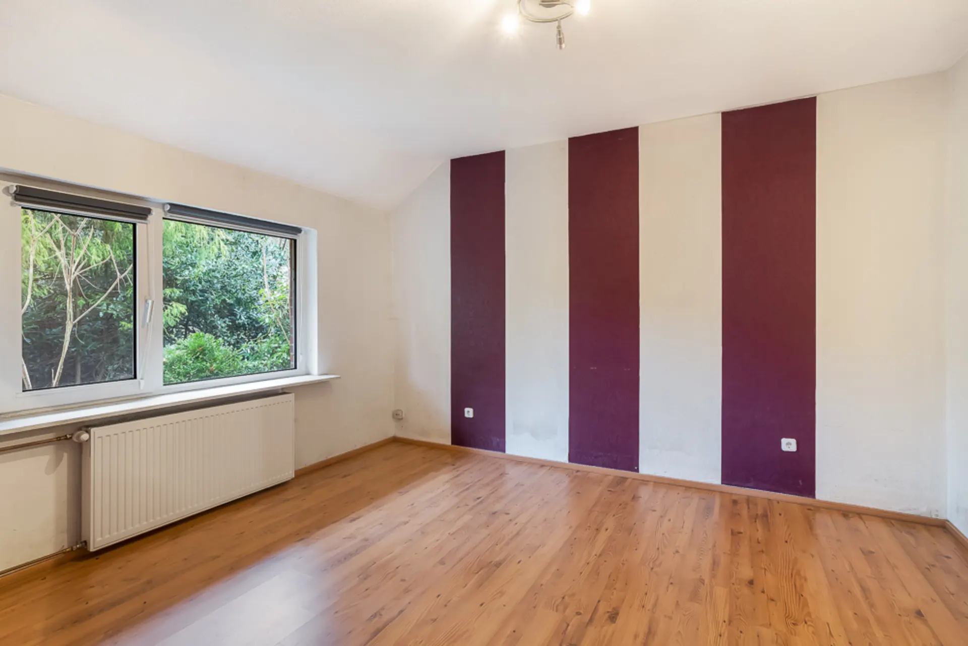 Leeres Zimmer mit Holzfußboden, großem Fenster und lila-weiß gestreifter Wand.