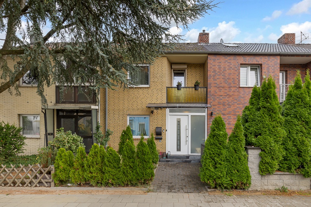 Zweistöckiges Wohnhaus mit Balkon, großer Eingangstür und begrüntem Vorgarten.