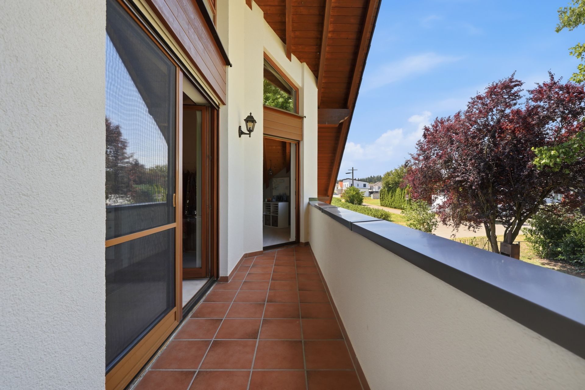Überdachter Balkon mit Terrakottafliesen, Blick auf Garten und Bäume, offene Glastür.