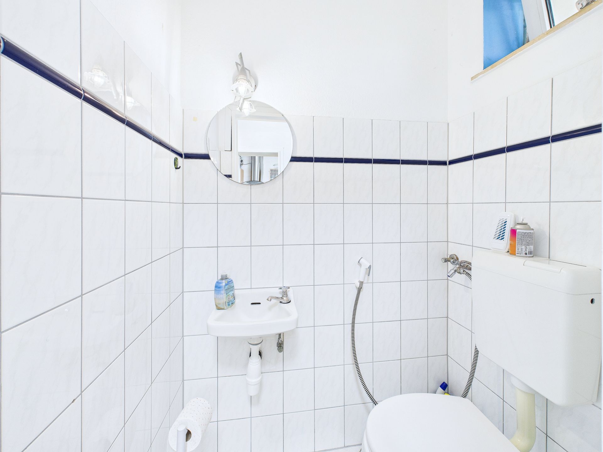 Kleines, weiß gefliestes Badezimmer mit WC, Waschbecken, Spiegel und Fenster.