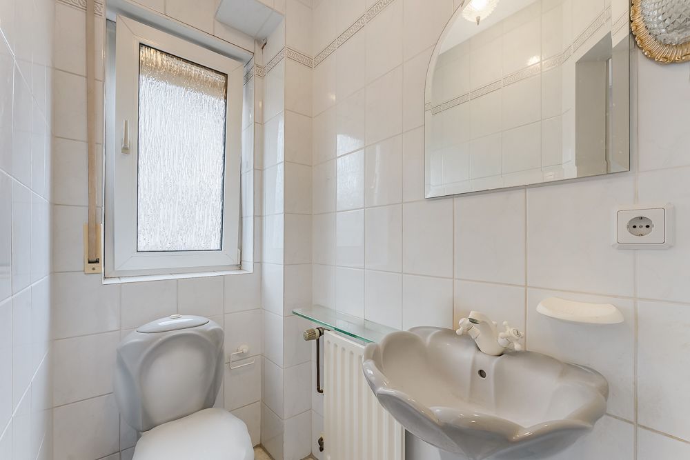 Kleines Badezimmer mit WC, Waschbecken, Spiegel und Fenster, weiße Fliesen an den Wänden.