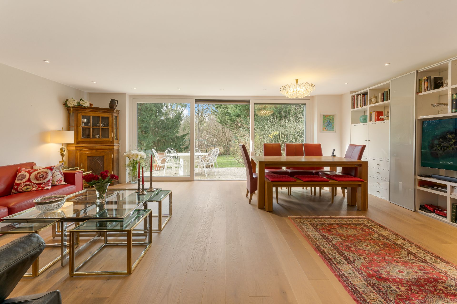 Helles Wohnzimmer mit rotem Sofa, Esstisch, Glastischen und Blick in den Garten.