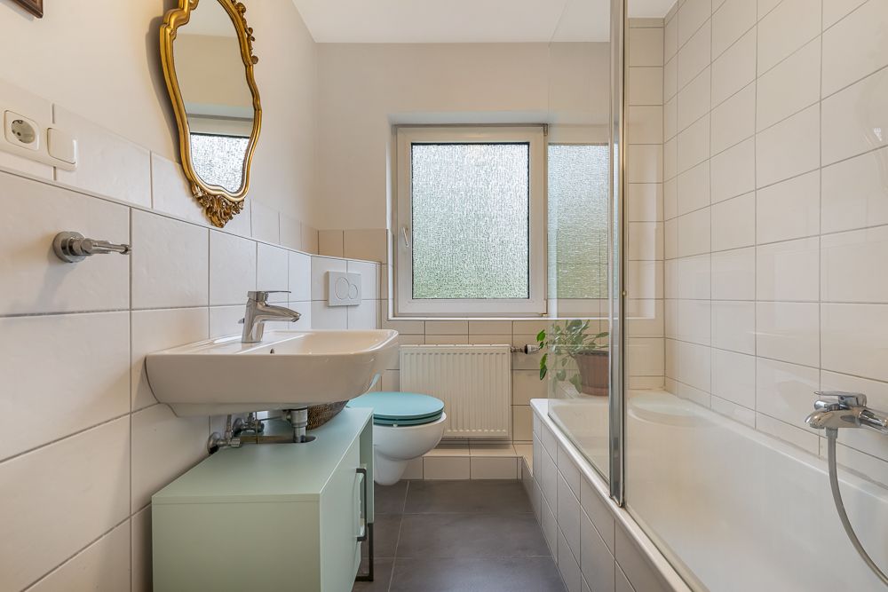 Helles Badezimmer mit Badewanne, Wandspiegel, Waschbecken, Toilette und Fenster.