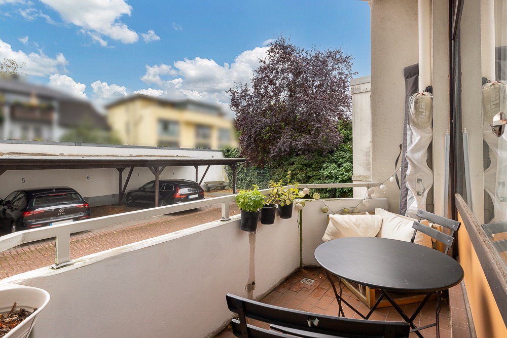 Kleine Balkonterrasse mit Tisch, Stühlen, Pflanzen und Blick auf Carport mit Autos.