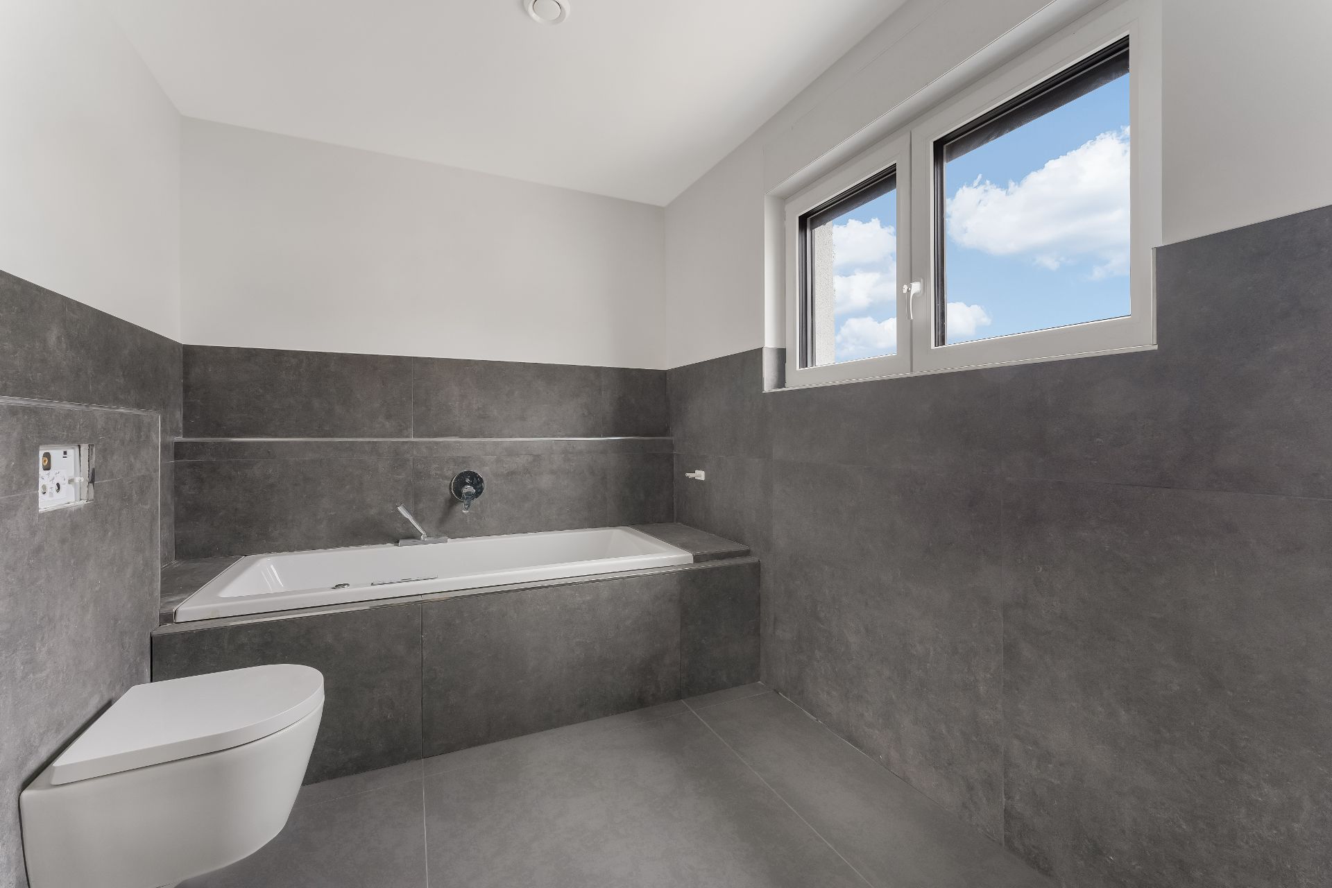 Modernes Badezimmer mit Badewanne, Wandtoilette und zwei Fenstern, graue Fliesen.
