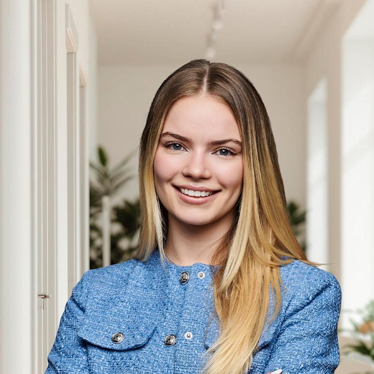 Junge Frau mit blauer Jacke steht mit verschränkten Armen in einem hellen Wohnraum.