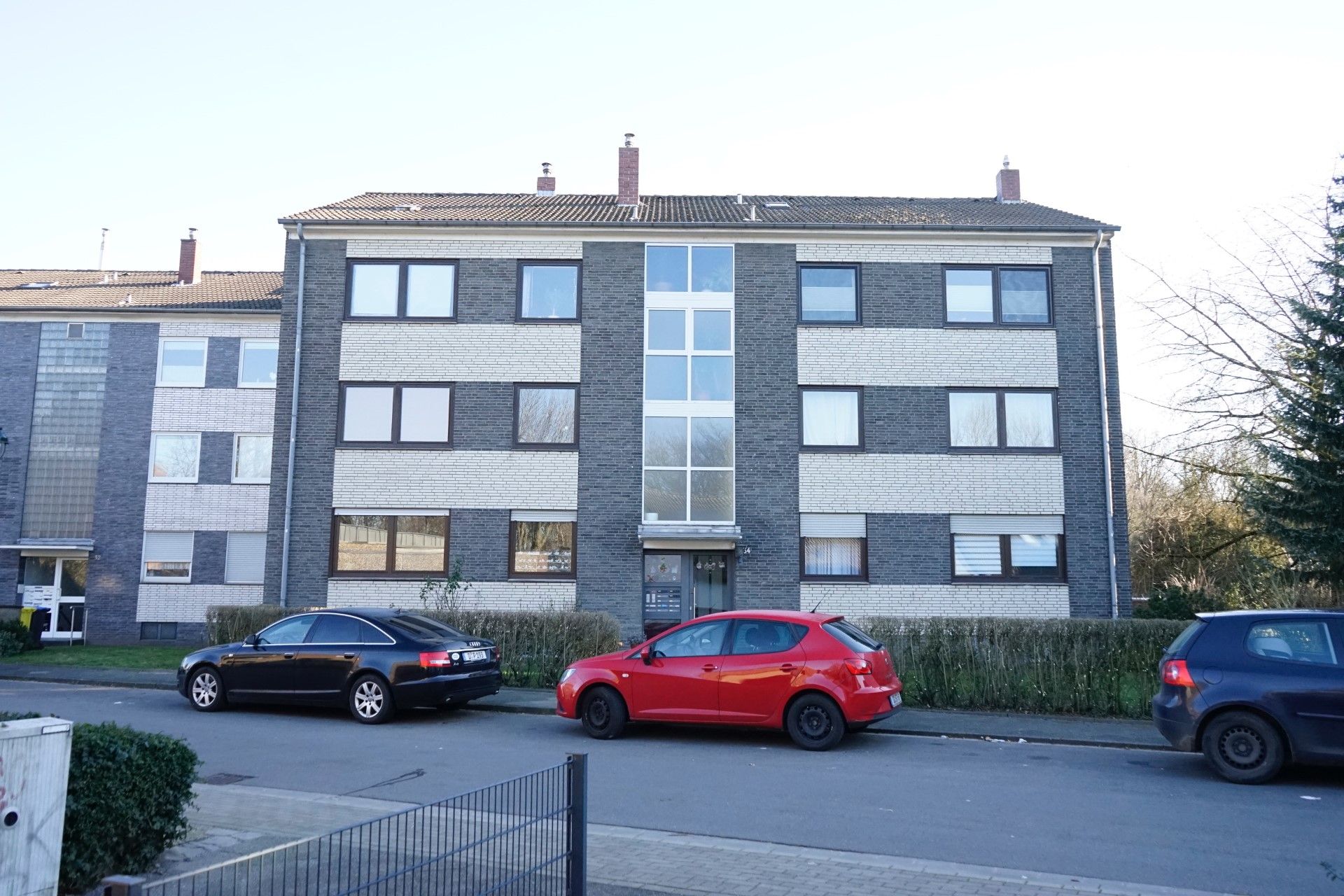 Mehrfamilienhaus mit drei Etagen und mehreren Fenstern, Autos parken davor auf der Straße.