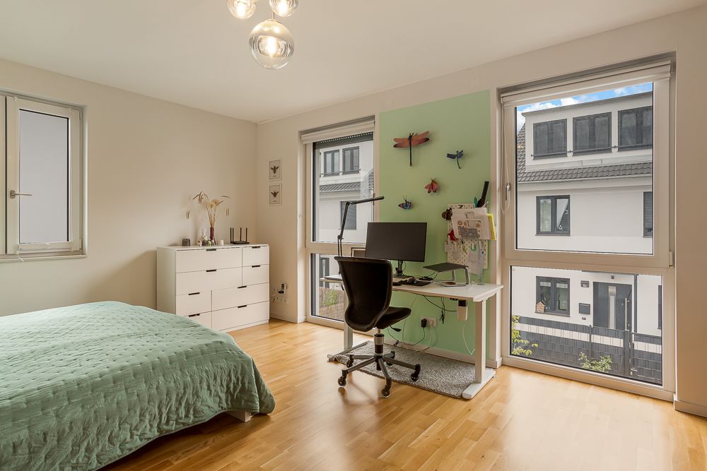 Helles Schlafzimmer mit Bett, Schreibtisch, Stuhl und Kommode, große Fenster mit Blick nach draußen.