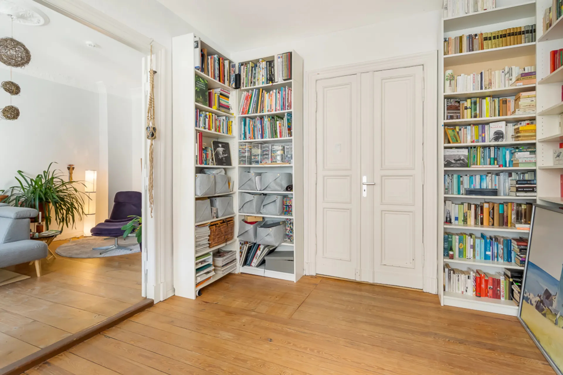 Heller Raum mit Bücherregalen, Holzboden und Blick in ein Wohnzimmer mit Pflanzen und Sessel.