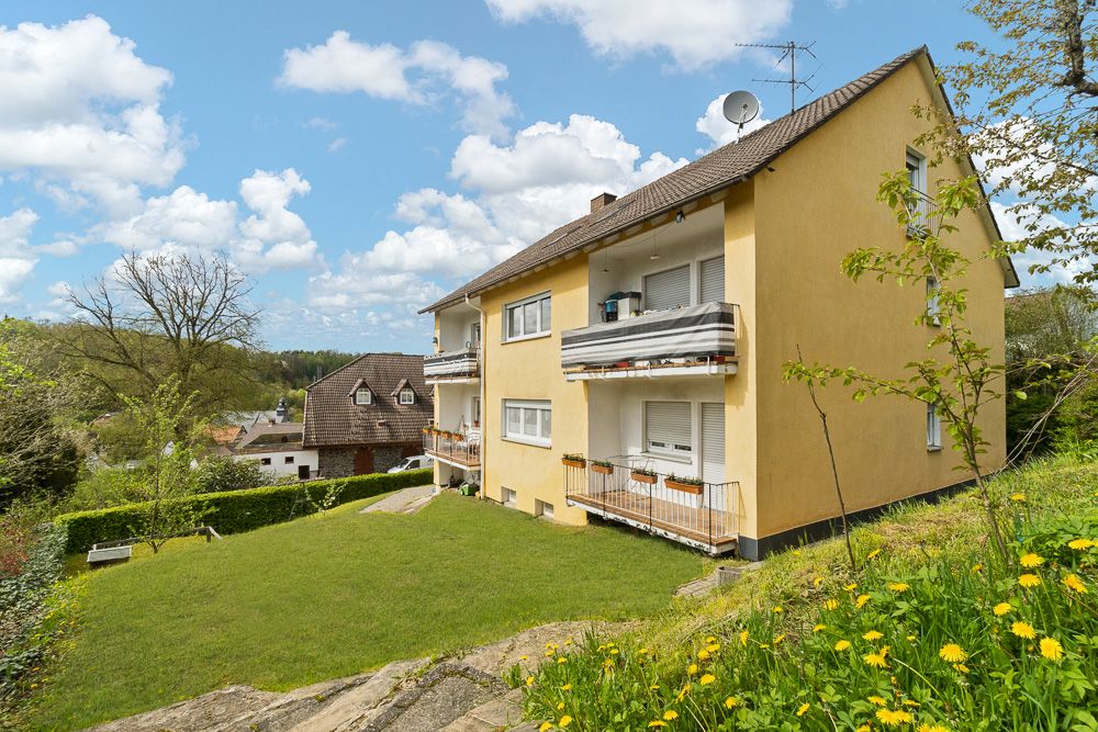 Gelbes Wohnhaus mit Balkonen und großem grünen Garten, umgeben von Bäumen und Nachbarhäusern.