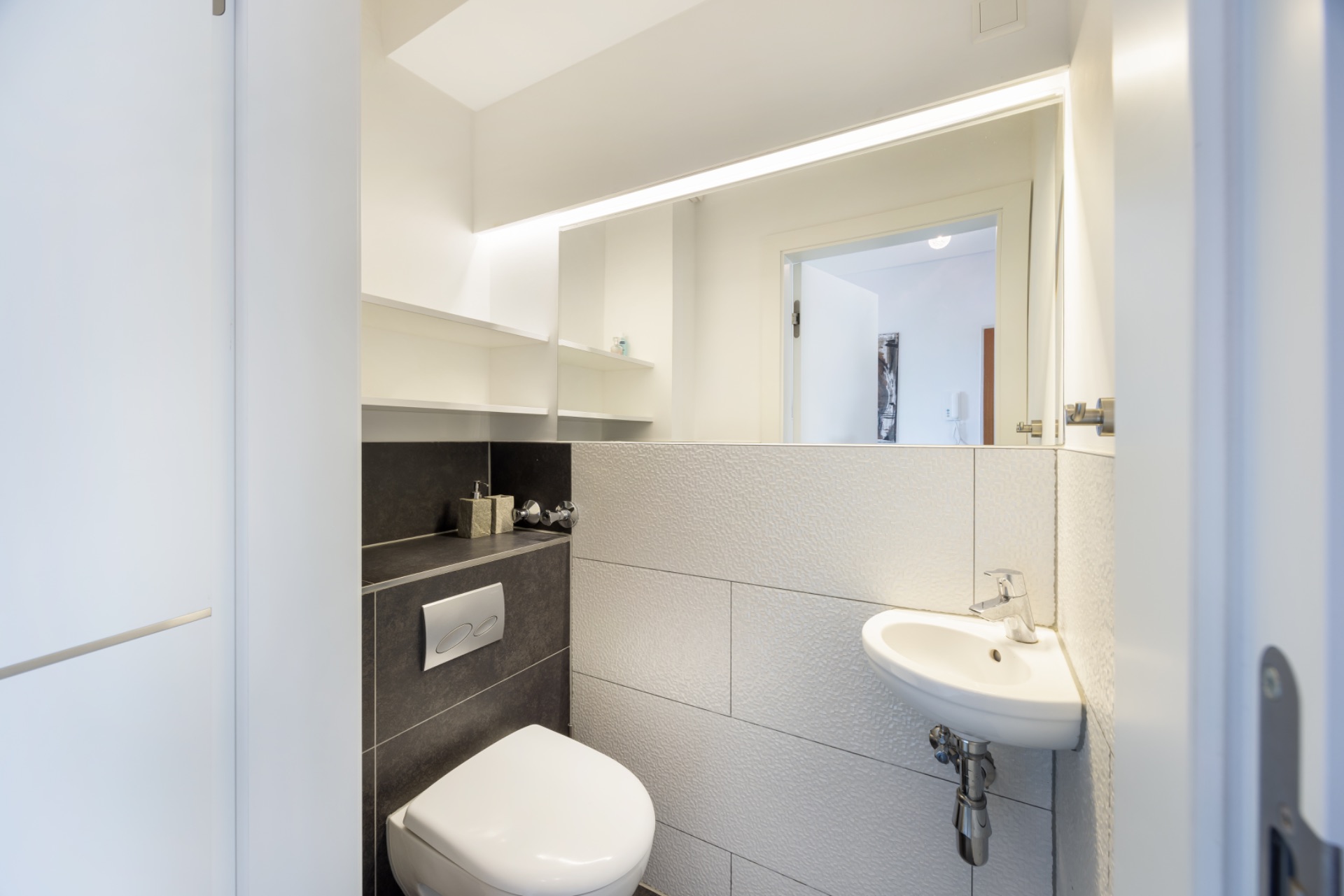 Kleines, modernes Bad mit Wandtoilette, kleinem Waschbecken und Spiegel.