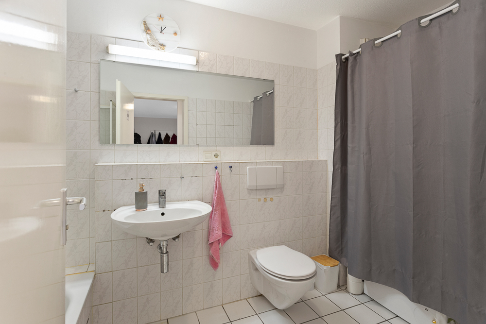 Helles Badezimmer mit Waschbecken, Spiegel, Toilette und Badewanne mit grauem Duschvorhang.