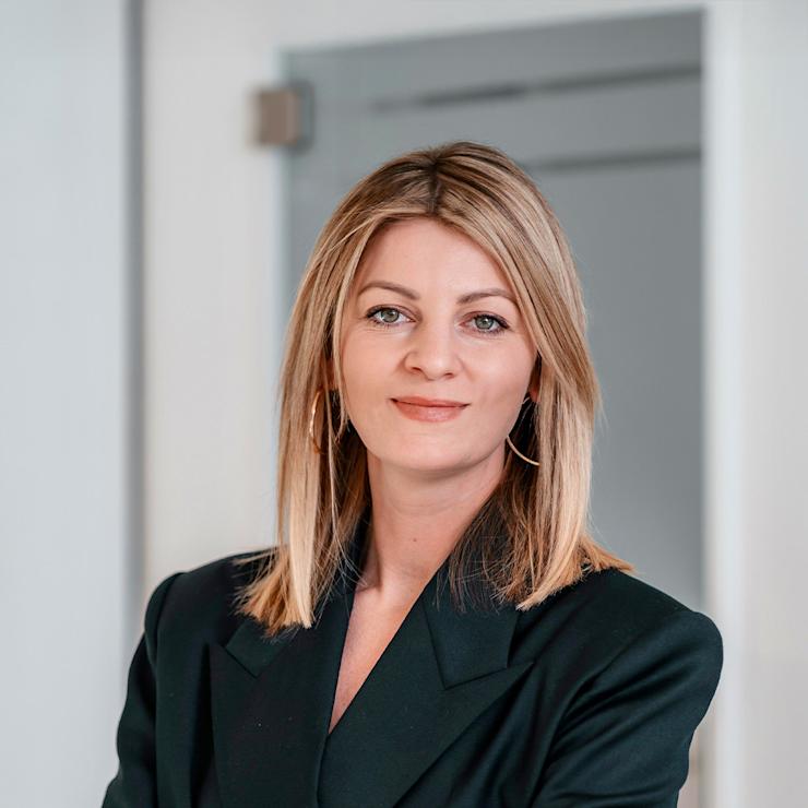 Frau mit blonden Haaren und schwarzem Blazer steht mit verschränkten Armen vor einer Tür.