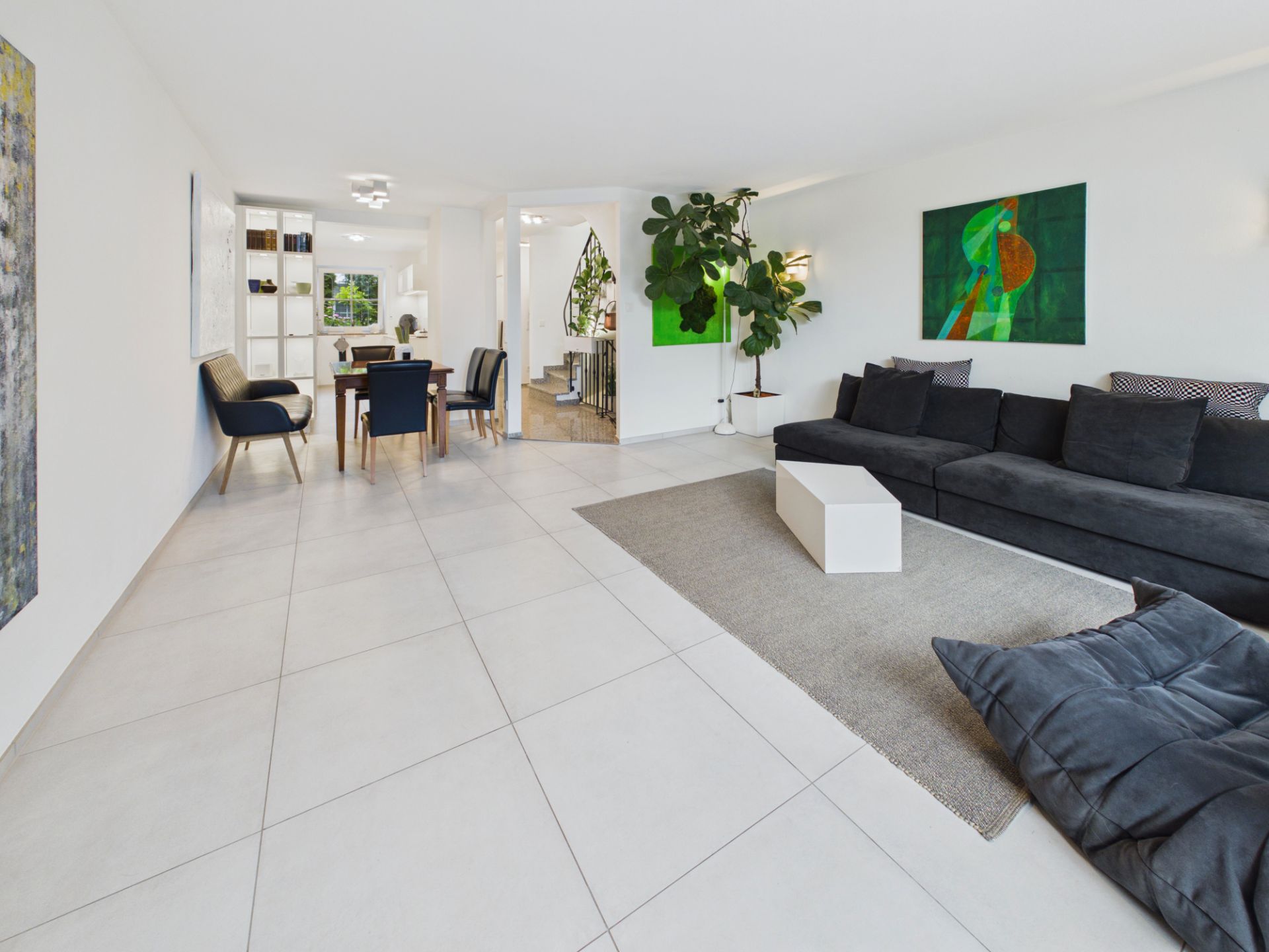 Helles Wohnzimmer mit großer dunkler Couch, Esstisch, Pflanzen und moderner Kunst an den Wänden.