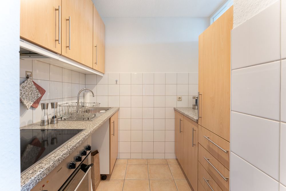 Schmale Küche mit Holzmöbeln, Elektroherd, Spüle und weiß gefliesten Wänden.