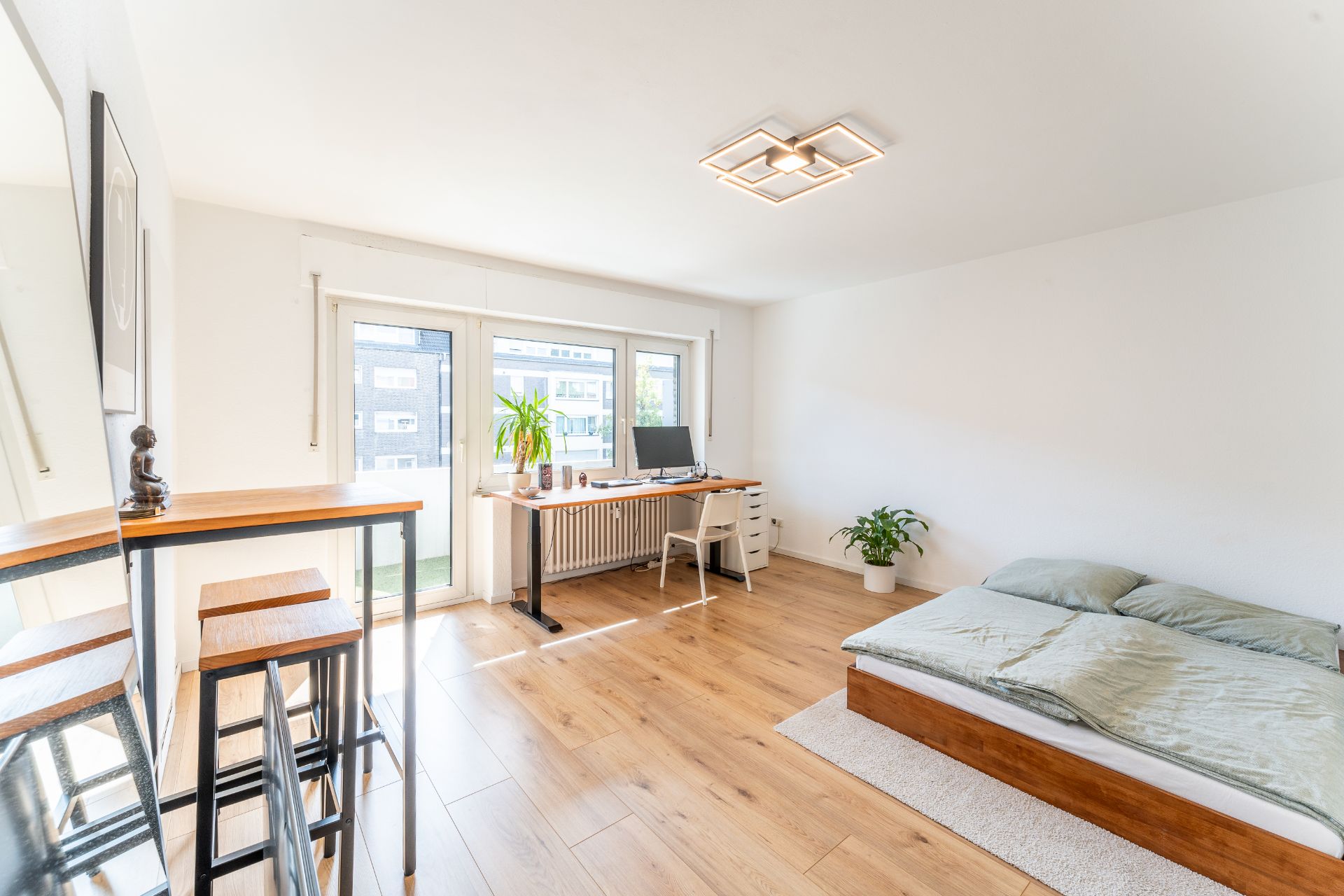 Helles Zimmer mit Bett, Schreibtisch, Barhockern und Pflanzen auf Holzboden.