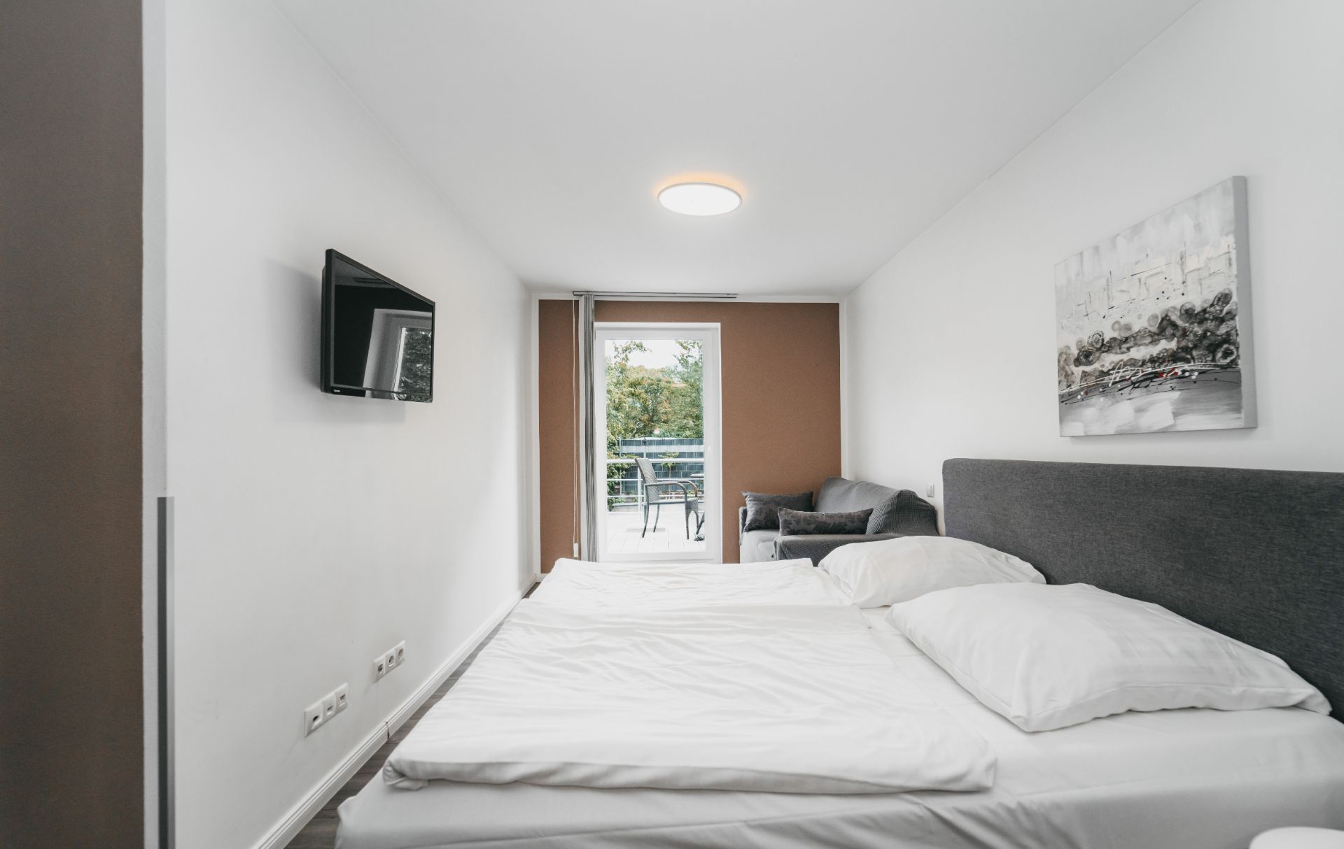 Helles Schlafzimmer mit Doppelbett, Sofa, Wandbild, TV und Zugang zu einem Balkon.