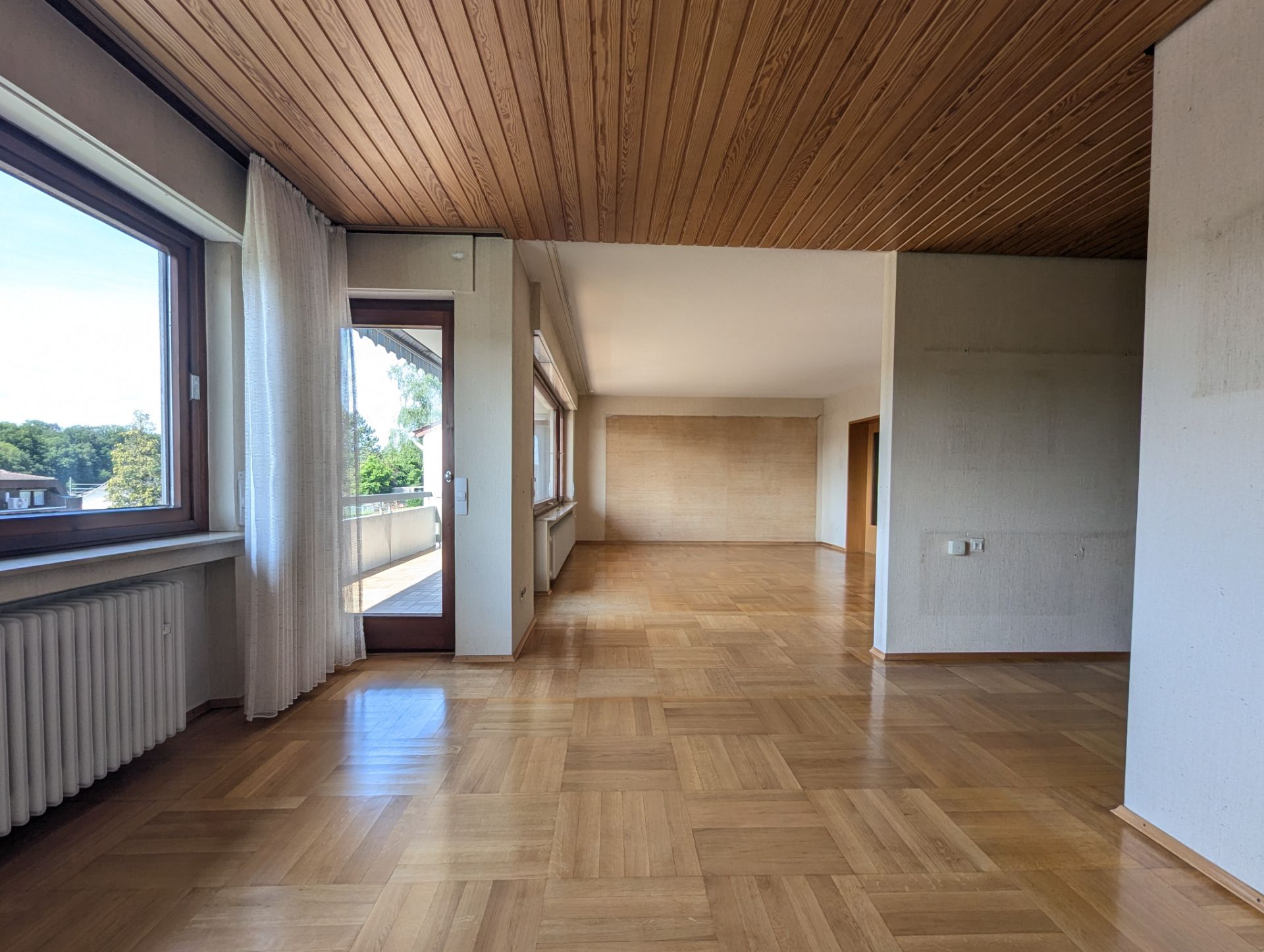Helles Wohnzimmer mit Parkettboden, Holzdecke und großen Fenstern. Zugang zum Balkon sichtbar.
