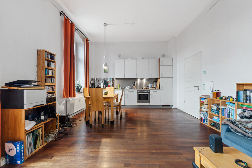 Helles Wohnzimmer mit offener Küche, Holzboden, Regalen und Esstisch mit vier Stühlen.