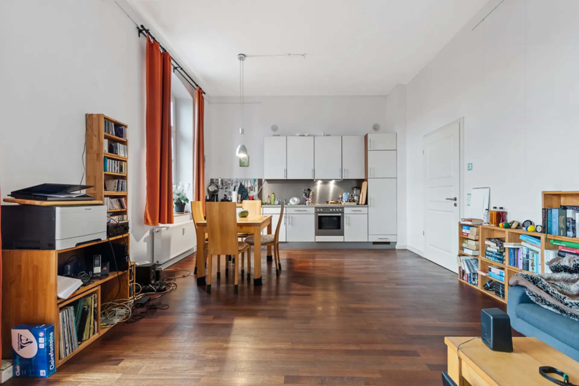 Helles Wohnzimmer mit offener Küche, Holzboden, Regalen und Esstisch mit vier Stühlen.