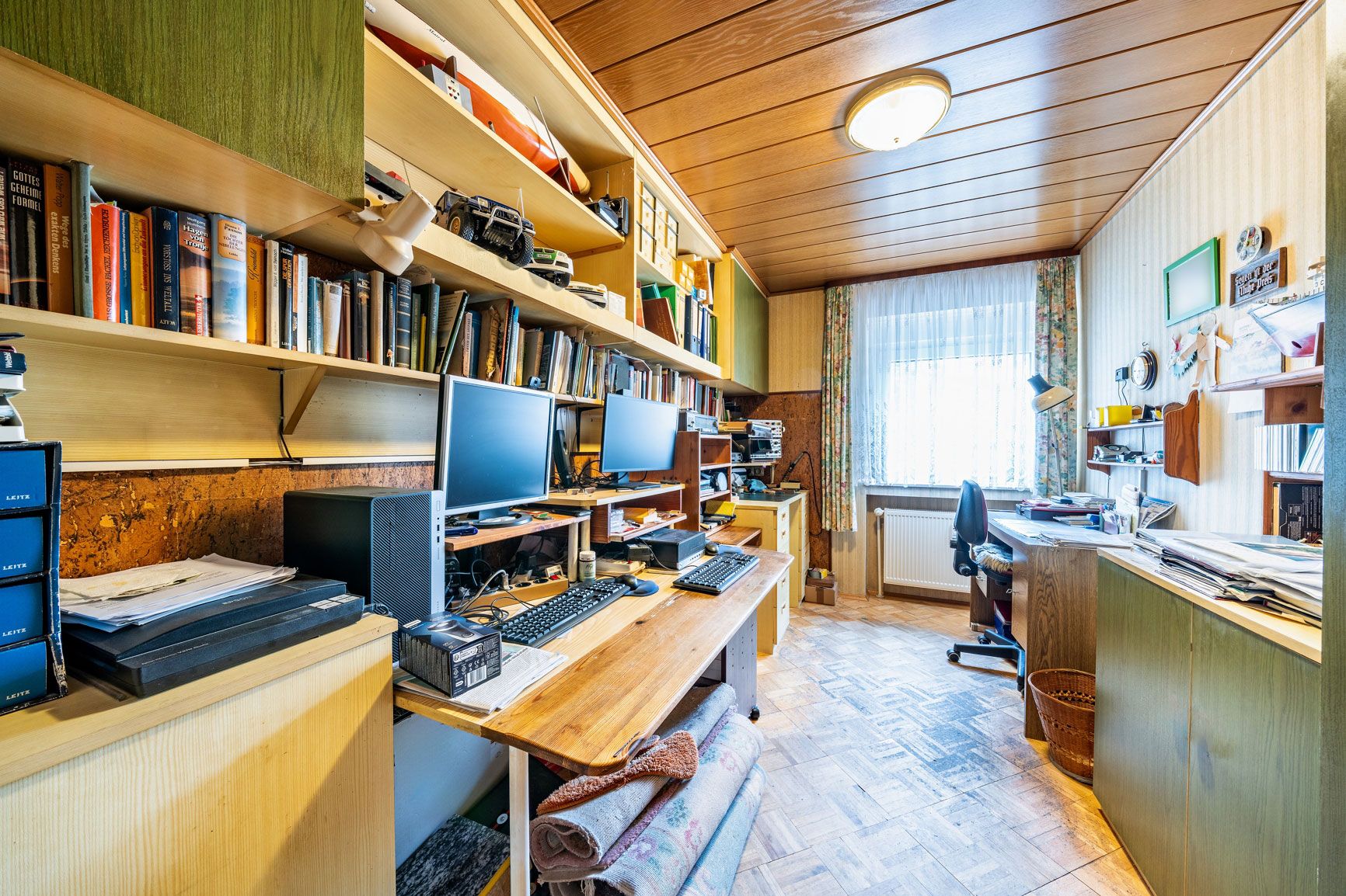 Arbeitszimmer mit zwei Schreibtischen, Regalen voller Bücher und zwei Monitoren, Fenster im Hintergrund.
