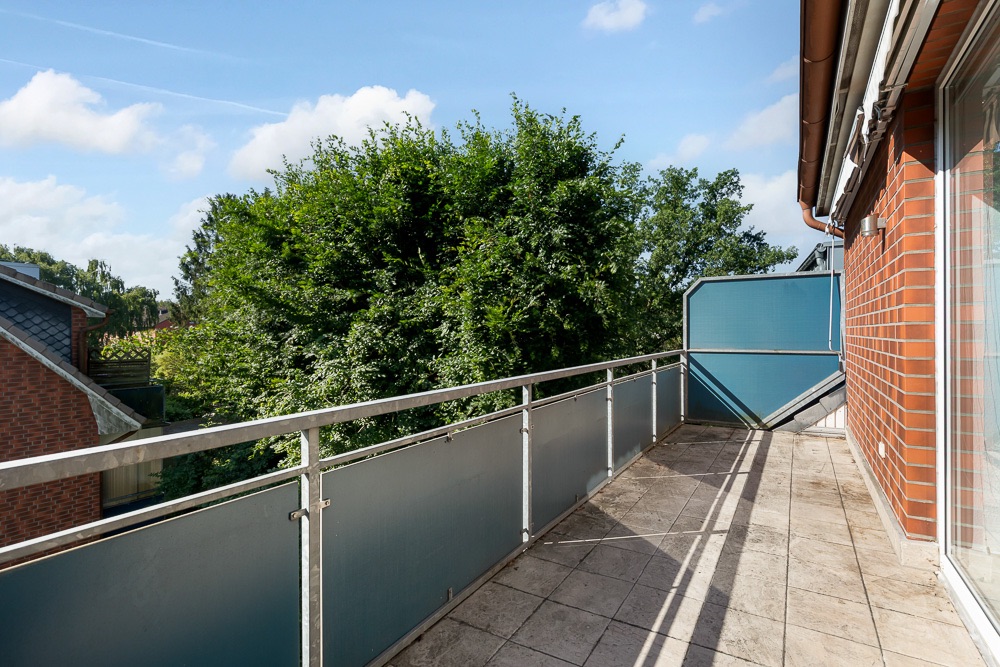 Balkon mit Fliesenboden und Metallgeländer, daneben rote Backsteinfassade, Blick auf Bäume.