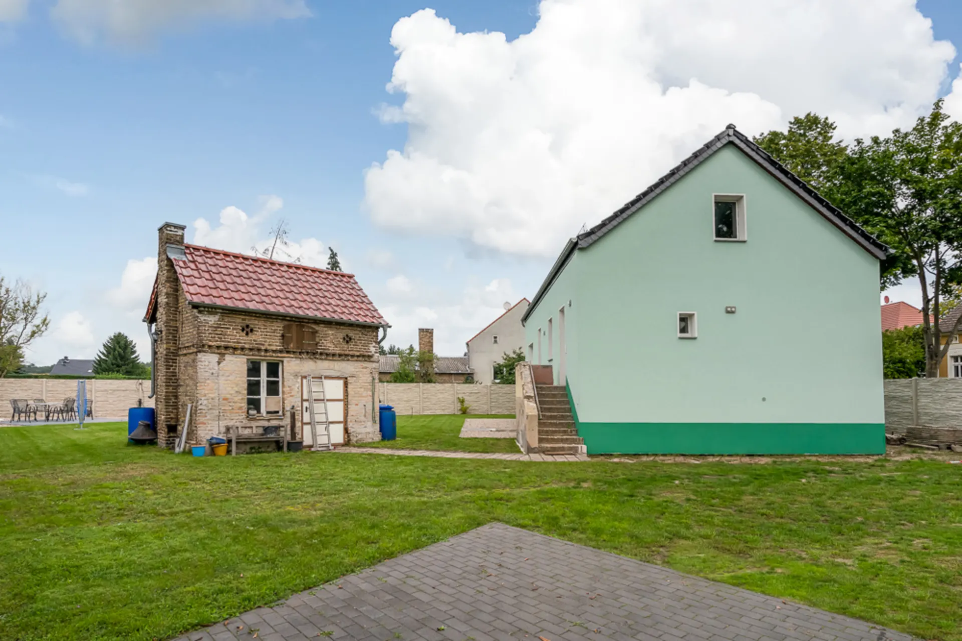 Zwei Häuser, eines alt aus Backstein mit rotem Dach, das andere grün gestrichen, auf einer Wiese.