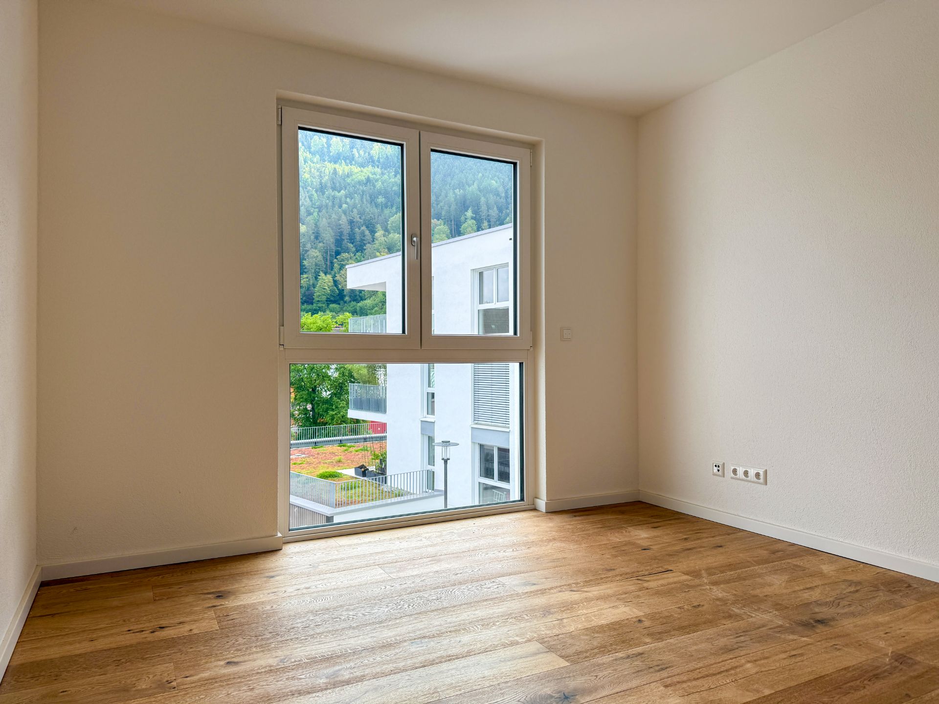 Leeres Zimmer mit Holzfußboden und großem Fenster, Blick auf modernes Wohngebäude und grüne Hügel.